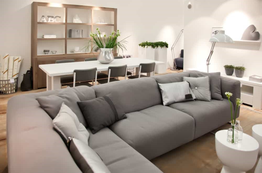 Ghế sofa xám lớn với gối tựa, bàn ăn trắng và kệ gỗ trong phòng khách hiện đại.